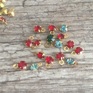 May include: Close-up of various small, colorful rhinestone charms with gold-colored loops. The charms feature red, green, and blue rhinestones. Some charms are clustered together, while others are scattered on a wooden surface.