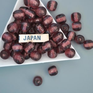 May include: A collection of dark red and black glass beads in a white triangular dish. A small rectangular sign with the word "JAPAN" is placed among the beads. The beads have a striped pattern and are likely used for jewelry making or crafts.