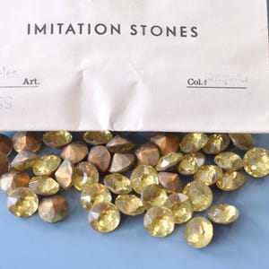 Puede incluir: Una colecci&oacute;n de piedras de imitaci&oacute;n en tonos dorados y amarillos. Algunas piedras tienen facetas, pareciendo gemas, mientras que otras son mate. Las piedras est&aacute;n dispersas sobre una superficie azul claro, con una etiqueta de papel que dice "IMITATION STONES".