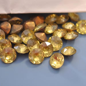 Puede incluir: Una colecci&oacute;n de pedrer&iacute;a facetada de color amarillo dorado y cuentas de color bronce con forma geom&eacute;trica. Los brillantes son redondos y reflejan la luz, mientras que las cuentas tienen un acabado mate. Los art&iacute;culos est&aacute;n dispersos sobre una superficie azul claro.