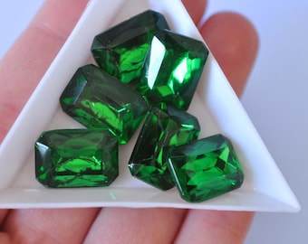 May include: A collection of emerald-cut, faceted green gemstones displayed in a white triangular tray. The gems have a vibrant, reflective quality, showcasing their polished surfaces and internal clarity. The tray is held in a person's hand.