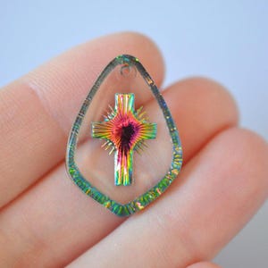 以下が含まれることがあります： カラフルな十字架のデザインが施された、透明な涙型のペンダント。十字架は赤、黄、緑、青など、鮮やかな色のスペクトルを特徴としています。ペンダントは手で持たれており、その虹色の特性を示しています。