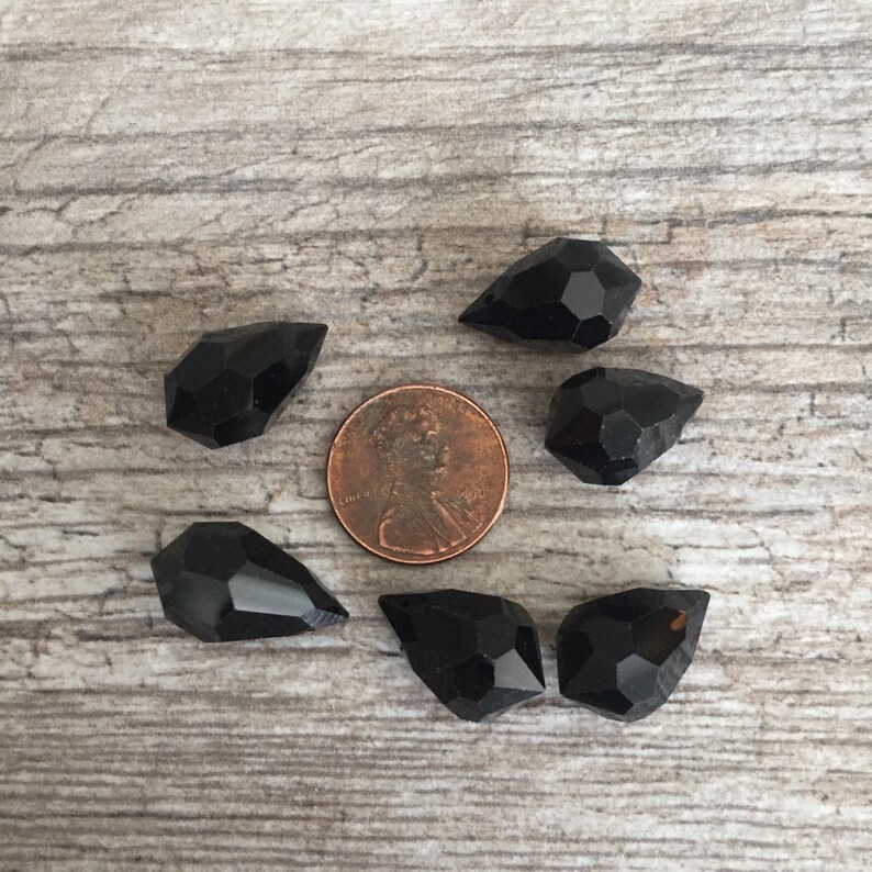 May include: Six black gemstone beads with a faceted design are arranged around a penny on a light brown wooden surface. The gemstones have a glossy finish and are various shapes.