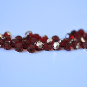 May include: A collection of faceted, deep red beads with metallic silver accents. The beads are scattered, showcasing their multiple reflective surfaces. The image is set against a soft, light blue background, highlighting the beads' color and texture.