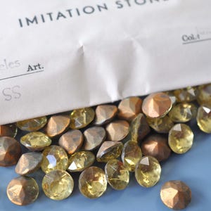 Puede incluir: Una colecci&oacute;n de piedras de imitaci&oacute;n en varias formas y colores, incluyendo gemas amarillas y bronce facetadas. Las piedras est&aacute;n dispersas sobre una superficie azul, con una bolsa de papel parcialmente visible etiquetada como "IMITATION STONE".