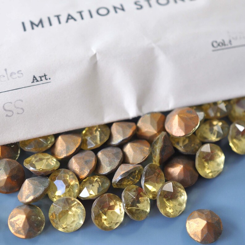 Puede incluir: Una colecci&oacute;n de piedras de imitaci&oacute;n en varias formas y colores, incluyendo gemas amarillas y bronce facetadas. Las piedras est&aacute;n dispersas sobre una superficie azul, con una bolsa de papel parcialmente visible etiquetada como "IMITATION STONE".