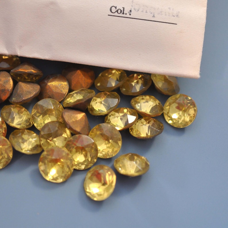 Puede incluir: Una colecci&oacute;n de gemas facetadas de color amarillo y piezas de color marr&oacute;n met&aacute;lico se encuentran dispersas sobre una superficie azul claro. Un trozo de papel con la palabra "Col." es parcialmente visible. Las gemas tienen talla brillante.