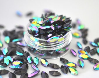 May include: A clear plastic jar filled with black, iridescent, marquise-shaped rhinestones. Some rhinestones are scattered around the jar on a white surface. The rhinestones have a rainbow sheen, reflecting colors like green, yellow, and purple.