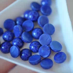 May include: A collection of round, blue lapis lazuli cabochons in various sizes, displayed on a white surface.
