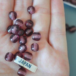 Op de afbeelding: Een verzameling kleine, doorschijnende, donkerpaarse kralen met een lichtere paarse streep, vastgehouden in een hand. Een klein label met het woord "JAPAN" is zichtbaar. Deze kralen worden waarschijnlijk gebruikt voor het maken van sieraden of knutselen.
