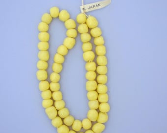 Op de afbeelding: Een ketting van lichtgele kralen met een enigszins onregelmatige vorm. De kralen zijn aan elkaar geregen en vormen een lange streng. Een klein label met de tekst "IN JAPAN" is aan de ketting bevestigd.