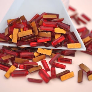 May include: A pile of red and gold rectangular glass rhinestones. The rhinestones are scattered on a pink surface.