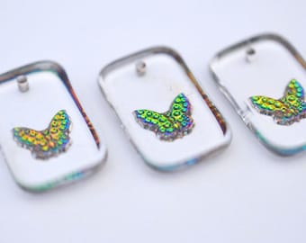 May include: Three clear, rectangular pendants featuring iridescent butterfly designs. Each pendant has a small hole at the top for hanging. The butterflies display a mix of green, yellow, and blue, creating a shimmering effect.