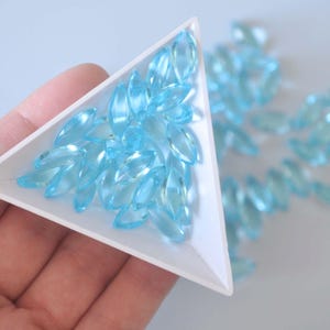 May include: A close-up of a triangular tray filled with light blue marquise-shaped gemstones. The tray is held in a hand.