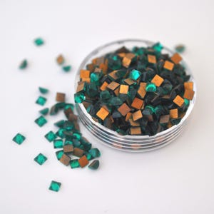 May include: A clear plastic container filled with small, square-shaped, emerald green and gold rhinestones. The rhinestones are scattered on a white surface.