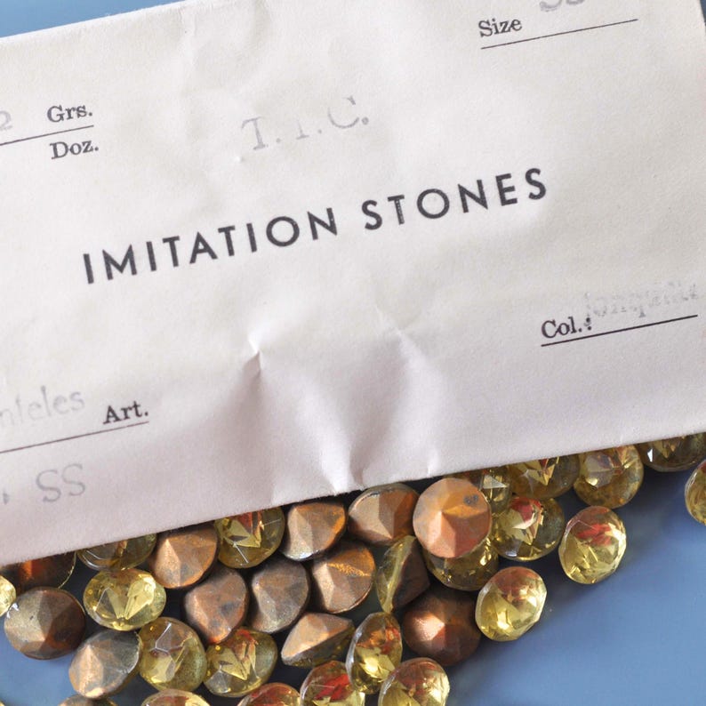 Puede incluir: Primer plano de piedras de imitaci&oacute;n doradas y marrones de varias formas. Las piedras est&aacute;n parcialmente cubiertas por un papel blanco con la inscripci&oacute;n "IMITATION STONES". Fondo azul.