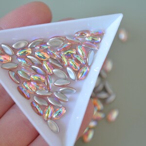 May include: A close-up of a white triangular tray filled with iridescent oval-shaped beads. The beads are a mix of silver and pink with an iridescent finish.