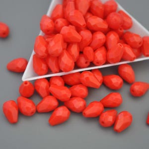 Könnte beinhalten: Helle, rote, facettierte Tropfenperlen. Die Perlen sind auf einer grauen Oberfläche verstreut, einige fallen aus einem weißen Behälter. Geeignet für Schmuckherstellung und Bastelprojekte.