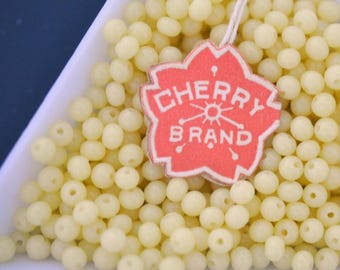 Op de afbeelding: Close-up van een witte bak gevuld met kleine, lichtgele kralen. Een rood label met de woorden "CHERRY BRAND" in witte letters is aan de kralen bevestigd. De kralen zijn uniform van grootte en vorm.