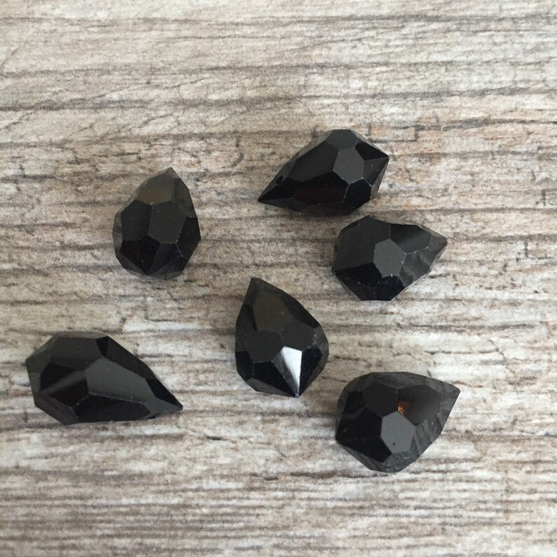 May include: Six black gemstone beads with faceted surfaces, in teardrop and other irregular shapes, are displayed on a light brown wooden background. The beads have a glossy finish, reflecting light and highlighting their geometric cuts.