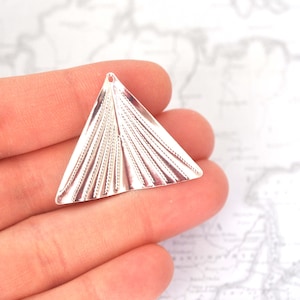 May include: A silver-colored, triangular pendant with a small hole at the top. The pendant features a fan-like design with etched lines radiating from the base. The pendant is held in a person's hand.