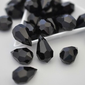 May include: Close-up of black faceted teardrop beads. The beads have a glossy finish and are scattered on a white surface. Some beads are in a white dish. The beads are a deep, solid black colour.