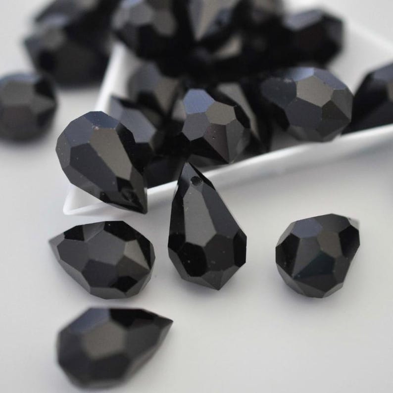 May include: Close-up of black faceted teardrop beads. The beads have a glossy finish and are scattered on a white surface. Some beads are in a white dish. The beads are a deep, solid black colour.