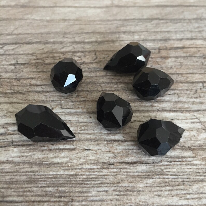 May include: Six black gemstone beads with multiple facets, in shapes like teardrops and rounded forms, are arranged on a light brown wooden surface. The beads have a glossy finish and reflect light.