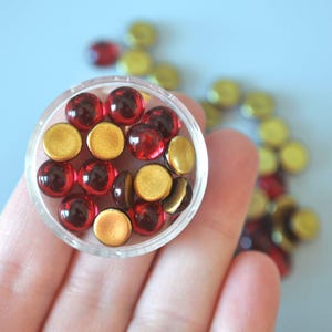 May include: A clear plastic container filled with red and gold round glass cabochons. The cabochons are arranged in a random pattern.