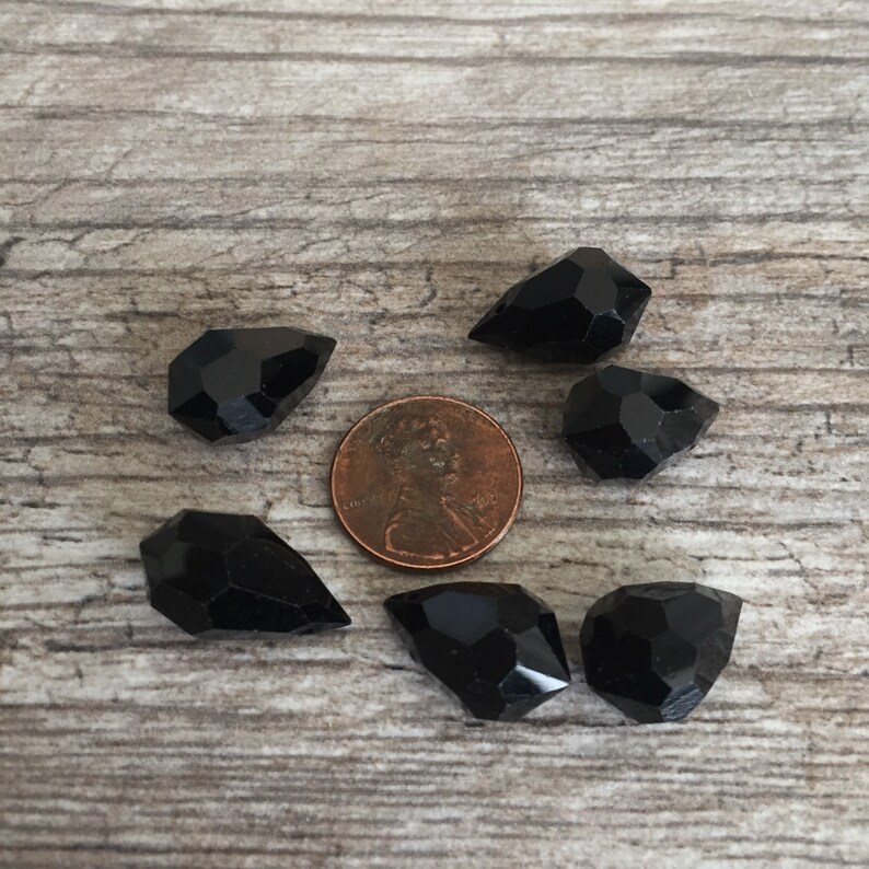 May include: Six black, faceted gemstone beads of different shapes are arranged around a penny on a wooden surface. The glossy beads reflect light. These beads are likely used for jewellery making or crafting.