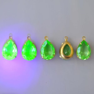 May include: Five teardrop-shaped pendants with gold-colored metal loops. Four pendants are bright green, faceted, and set in gold-colored metal. The fifth pendant has a gold-colored metal frame and a green stone. The pendants are displayed on a gray background.