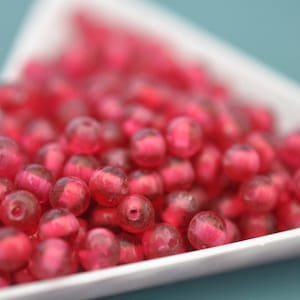 May include: Close-up of a pile of translucent, round, red beads. The beads are clustered together in a white dish, with a blurred background. The beads have a glossy finish and appear to be made of glass or a similar material. Ideal for jewelry making.
