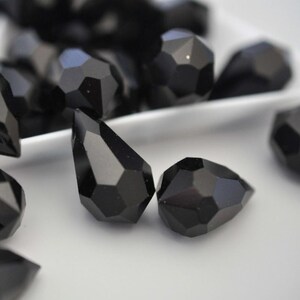 May include: Close-up of several faceted black gemstone beads. The beads have a geometric, multi-faceted design, with a teardrop shape and a round shape. The beads are displayed on a white surface, with some beads in the background.