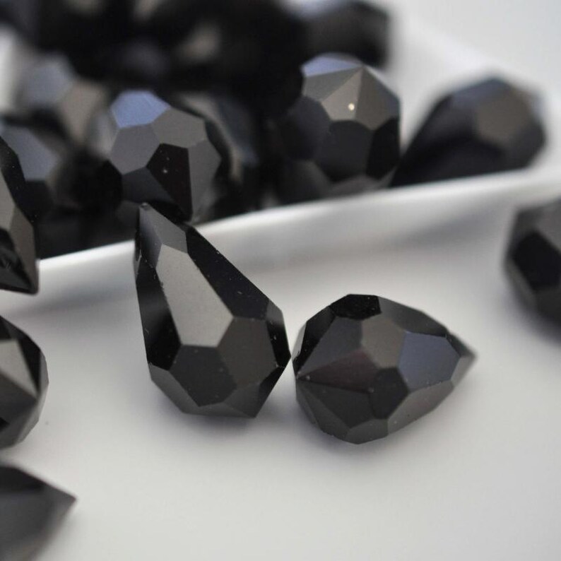 May include: Close-up of several faceted black gemstone beads. The beads have a geometric, multi-faceted design, with a teardrop shape and a round shape. The beads are displayed on a white surface, with some beads in the background.