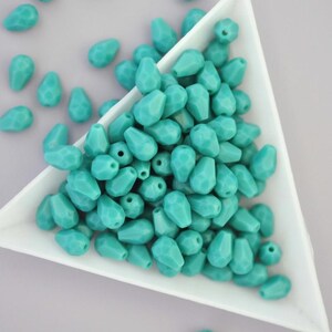 May include: A collection of turquoise teardrop-shaped beads with faceted surfaces. The beads are scattered in a white triangular dish and on a light grey surface. These beads are suitable for jewellery making and crafting projects.