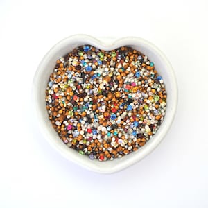 May include: A white, heart-shaped ceramic bowl filled with a mix of colourful rhinestones. The rhinestones are in shades of gold, silver, red, blue, green, and iridescent colours. The bowl is on a white background.