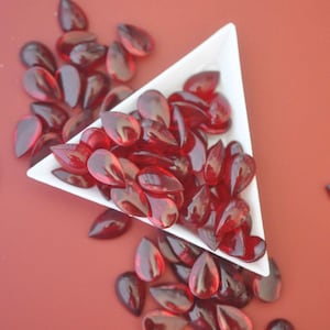 May include: A white triangular dish filled with red teardrop-shaped glass beads. The dish is on a red surface and there are more beads scattered around it.