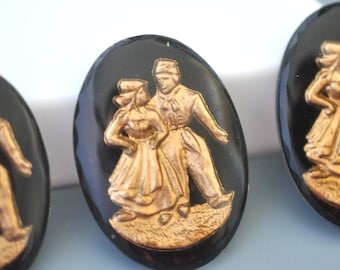 May include: Two gold-coloured figures of a couple dancing, set against a black oval background. The figures are raised and have a 3D effect.