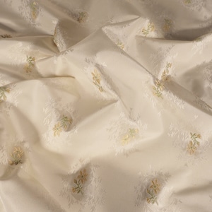 Puede incluir: Primer plano de una tela blanca con un sutil estampado floral en tonos verdes y amarillos. La tela es suave y tiene una superficie ligeramente texturizada.