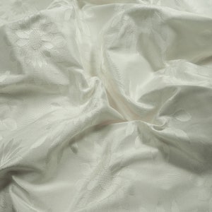 Könnte beinhalten: Weißer Stoff mit einem erhabenen Blumenmuster. Der Stoff ist zerknittert und hat ein weiches, luxuriöses Gefühl.