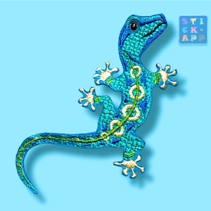Könnte beinhalten: Gestickter Aufnäher eines türkisfarbenen Geckos mit weißen und goldenen Akzenten. Der Gecko hat ein detailliertes Design mit einem grünen Rückgrat und ist vor einem hellblauen Hintergrund platziert. Der Aufnäher enthält den Text "STICK APP".