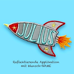 Könnte beinhalten: Eine silberne und rote Rakete mit einem blauen gestickten Namen "JULIUS" und gelben Sternen. Die Rakete hat eine Flamme, die aus dem Heck kommt. Der Text "Reflektierende Applikation mit Wunsch-NAME" befindet sich unter der Rakete.