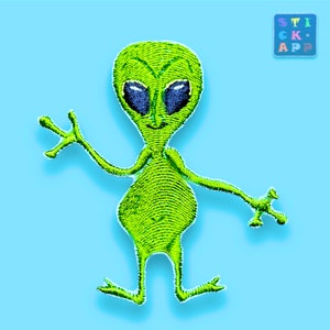 Könnte beinhalten: Gestickter grüner Alien-Aufnäher mit großen blauen Augen und winkenden Armen, vor hellblauem Hintergrund. Der Alien hat einen lächelnden Ausdruck und ein strukturiertes, genähtes Aussehen. Der Text "STICK APP" befindet sich in der oberen rechten Ecke.