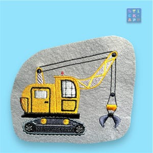 Könnte beinhalten: Gelber gestickter Baukran mit schwarzer Raupe und grauem Greifarm. Der Kran befindet sich auf einem grauen Filz-Hintergrund.