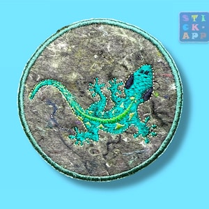 Könnte beinhalten: Runder, gestickter Aufnäher mit einem türkisfarbenen Eidechsen-Design, schwarzen Punkten und gelben Akzenten auf einem strukturierten grauen Hintergrund. Der Aufnäher hat einen grünen Rand. Der Text "STICK APP" ist oben rechts zu sehen.