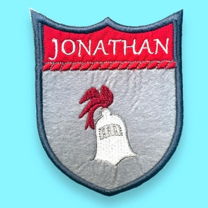 Könnte beinhalten: Ein grauer Filzschild mit einem roten und weißen Ritterhelm-Design. Der Schild hat oben ein rotes Banner mit dem Namen "JONATHAN" in weißen Buchstaben.