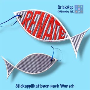 Könnte beinhalten: Zwei fischförmige Stoffanhänger mit einem marineblau-weißen Nadelstreifenmuster. Der größere Fisch zeigt den Namen RENATE in kräftigem rotem Garn gestickt. Jeder Anhänger besitzt eine kleine Metallöse und ein schmales blaues Band zum Aufhängen.