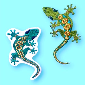 Könnte beinhalten: Zwei gestickte Bügelbilder von grünen Geckos mit goldenen und blauen Details. Die Geckos sind in einer zusammengerollten Position mit ihren Schwänzen um ihre Körper gewickelt.