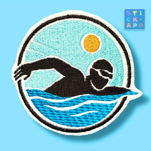 Könnte beinhalten: Ein runder gestickter Aufnäher mit der schwarzen Silhouette einer schwimmenden Person in blauem Wasser mit weißen Wellendetails. Der Hintergrund ist hellblau mit einer gelben Sonne in der oberen rechten Ecke, umrandet von einem schwarzen Rand.