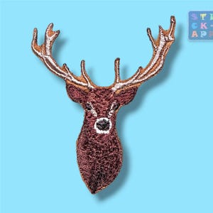 Könnte beinhalten: Ein gestickter Aufnäher, der einen Hirschkopf mit großem, detailliertem Geweih zeigt. Der Hirsch ist in Brauntönen mit einer weißen Schnauze und dunklen Augen dargestellt. Die Stickerei weist ein strukturiertes Finish auf, das die Merkmale des Tieres vor einem einfarbigen hellblauen Hintergrund hervorhebt.